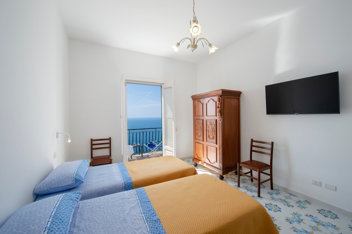 A twin room is presented with two single beds, featuring soft blue and yellow linens. A large window offers a view of the sea and leads to a balcony, while a wooden wardrobe and a chair complement the space. Natural light brightens the room.