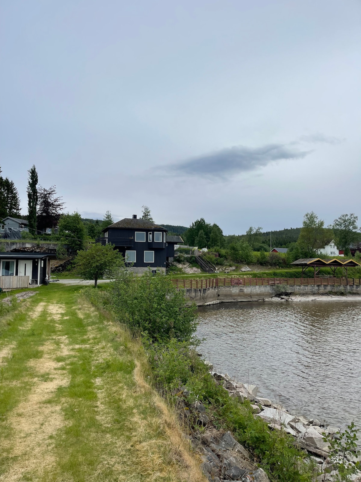 A well-maintained grassy path runs alongside a calm body of water, surrounded by trees and shrubs. A dark-colored house is visible in the background, with additional structures partially obscured by greenery. The landscape features a gentle slope leading down to the water's edge.