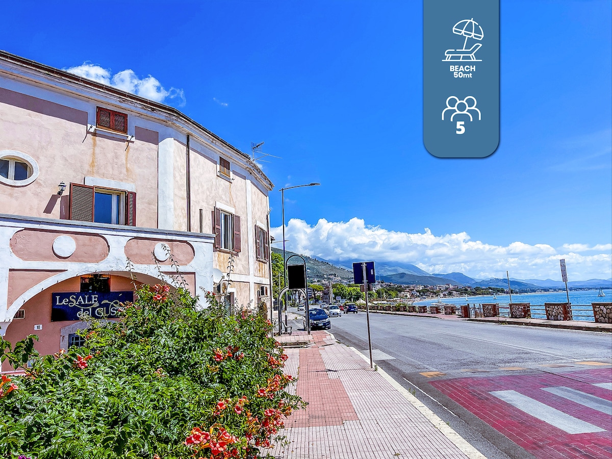 The image depicts a coastal street with a pastel-colored building featuring large windows. Lush greenery and vibrant flowers line the sidewalk, leading towards the beach in the distance. Bright blue skies with scattered clouds create an inviting atmosphere along the waterfront.