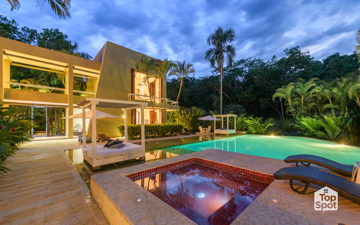 The two-story house is surrounded by lush greenery, featuring a tranquil swimming pool and a jacuzzi. Comfortable lounge chairs are positioned near the water, creating a serene outdoor space. The evening sky is adorned with soft clouds, enhancing the peaceful ambiance.