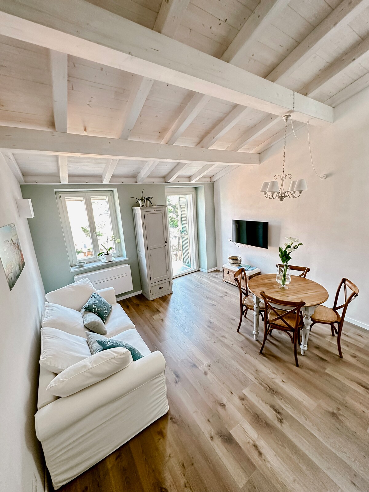 The living area features a cozy white sofa, a round wooden dining table surrounded by four wooden chairs, and a flat-screen television mounted on the wall. Natural light streams in from two windows, enhancing the open feel of the space with light-colored walls and a wooden ceiling.