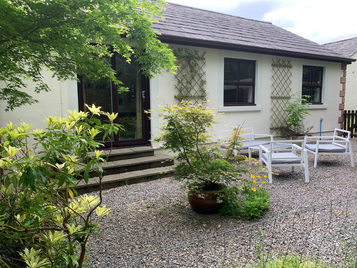 The exterior of the cottage is framed by lush greenery and flowering plants. A pebbled area features white outdoor furniture arranged for relaxation. Climbing plants adorn the trellis beside large windows, inviting natural light into the space.