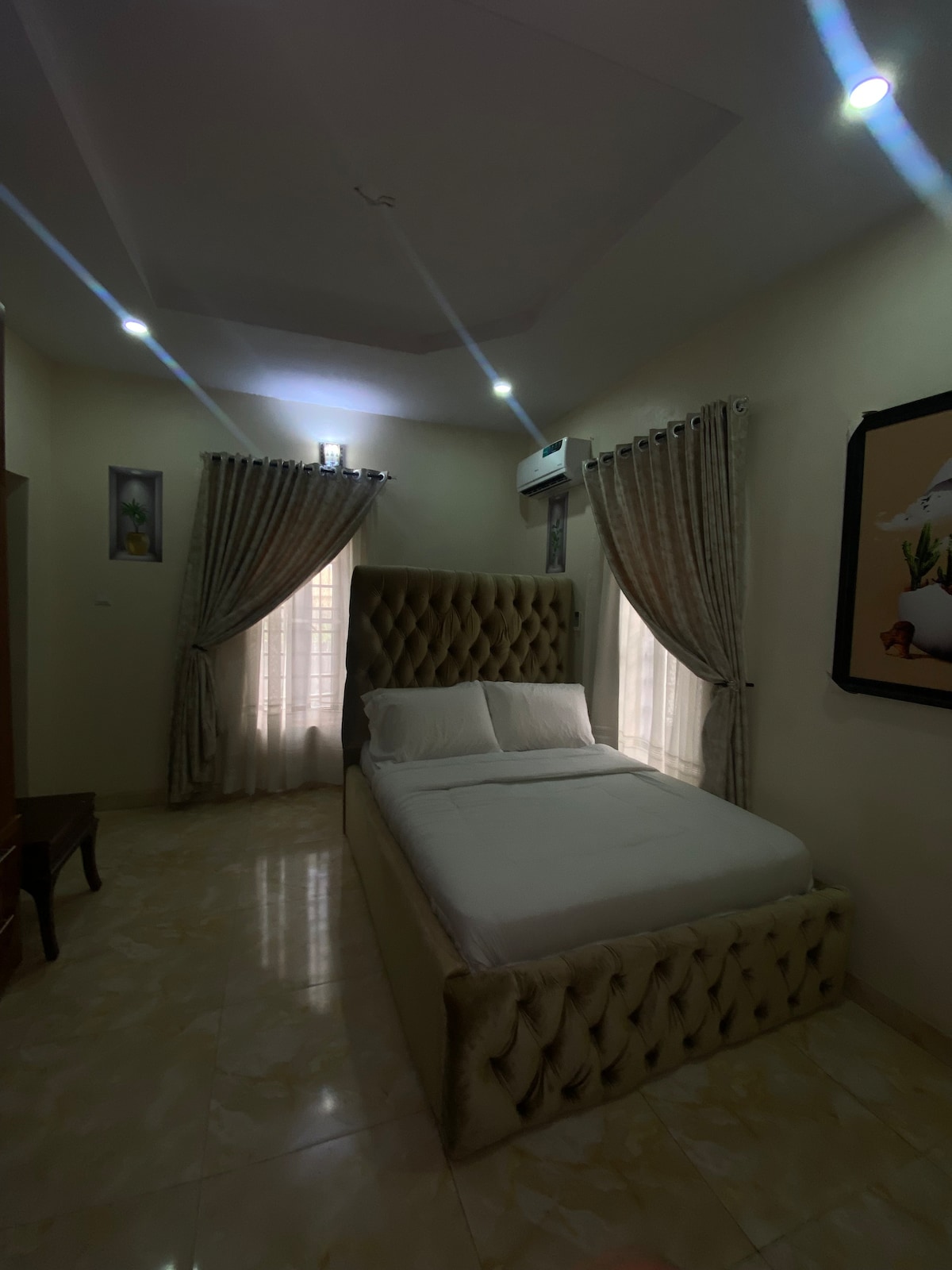 A well-appointed sleeping area features a spacious bed with a padded headboard, complemented by soft bedding. Natural light filters through two window drapes, creating a serene environment. Walls are adorned with framed artwork, and an air conditioning unit is mounted for comfort.