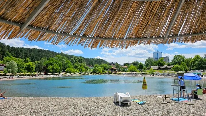 A scenic view of a pebbled beach area beside a calm body of water, framed by a thatched roof. Sun loungers are positioned on the shore, while umbrellas provide shade. Lush greenery and scattered visitors can be seen in the background, along with distant structures.