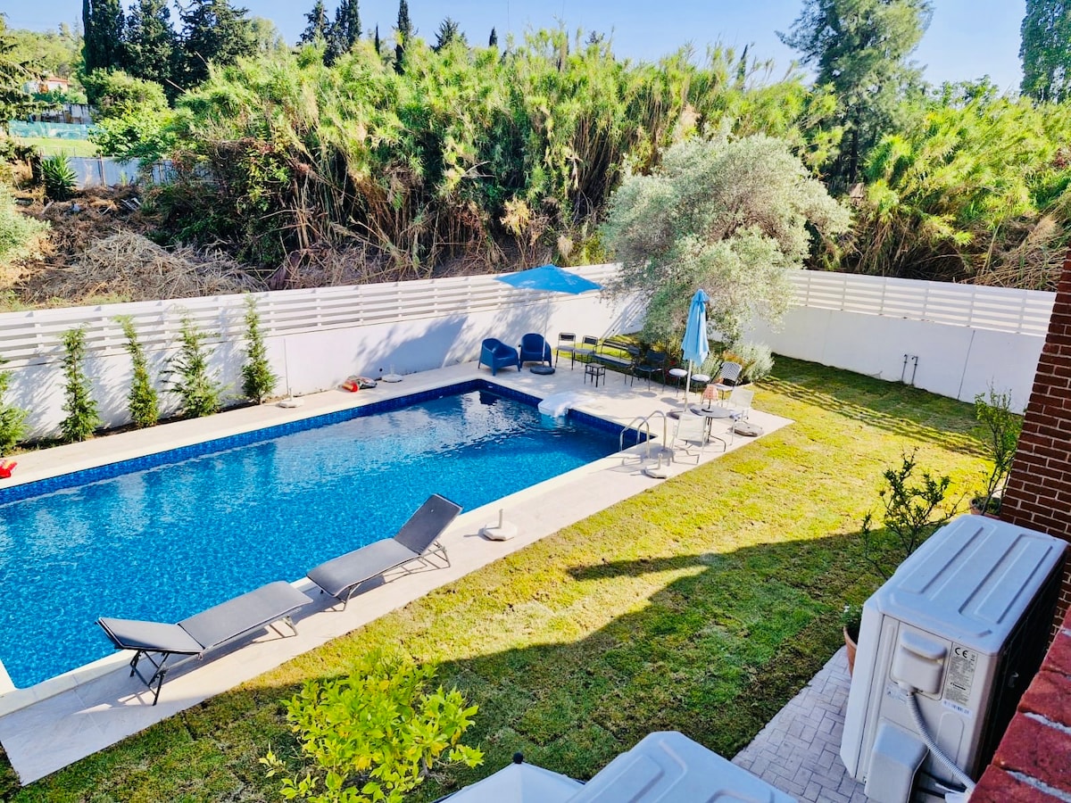The outdoor area features a swimming pool surrounded by landscaped greenery. Sun loungers are positioned on a deck beside the pool, with a shaded seating area and an umbrella providing cool relief. A manicured lawn complements the inviting space.
