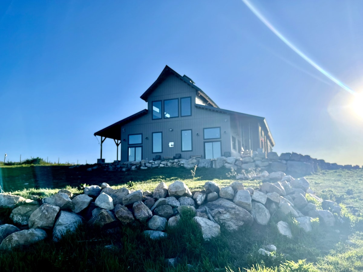 A barn-style cabin is positioned on a slight elevation, surrounded by a landscaped area featuring stones. The exterior showcases large windows, and the sloped roof adds architectural interest against the clear blue sky.