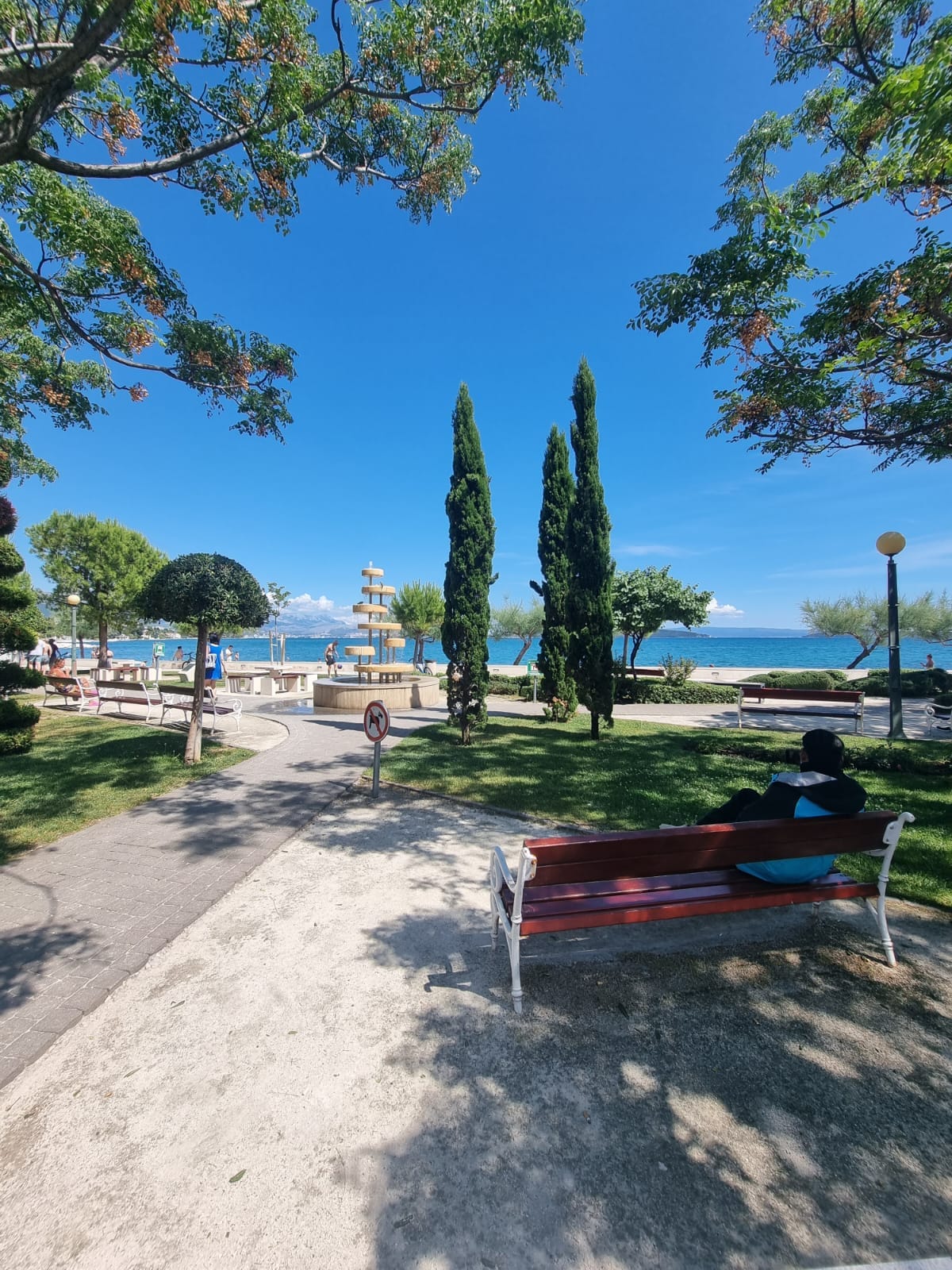 A well-maintained park pathway is lined with leafy trees and manicured grass. A red bench provides a seating area along the path, facing a view of the sea and nearby structures. Bright blue skies enhance the inviting outdoor space.