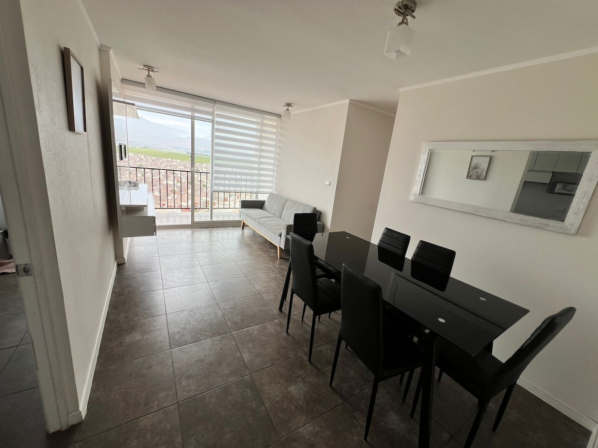 An open living and dining area is captured, featuring a sleek dining table with black chairs and a light-colored sofa positioned near large windows. Natural light fills the space, highlighting the neutral color palette and modern furnishings.