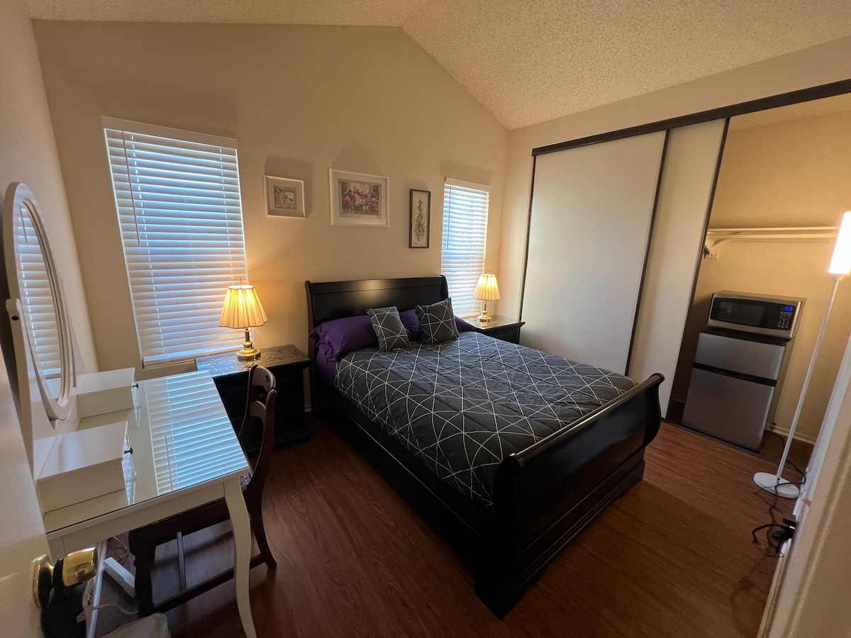 A cozy second-story room contains a full-size bed with dark bedding and purple accent pillows. A desk and chair are positioned by a window, while a refrigerator and microwave are located in a closet. Two windows allow natural light to brighten the space.