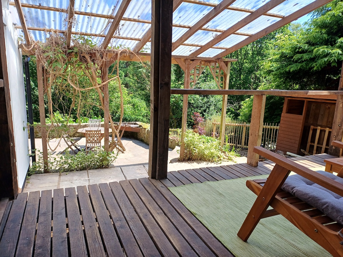 An outdoor patio area features wooden decking and a covered section with clear roofing. Greenery surrounds the space, with plants climbing the support beams. A comfortable lounge chair and a small table are positioned in a sunlit corner, overlooking the peaceful garden.