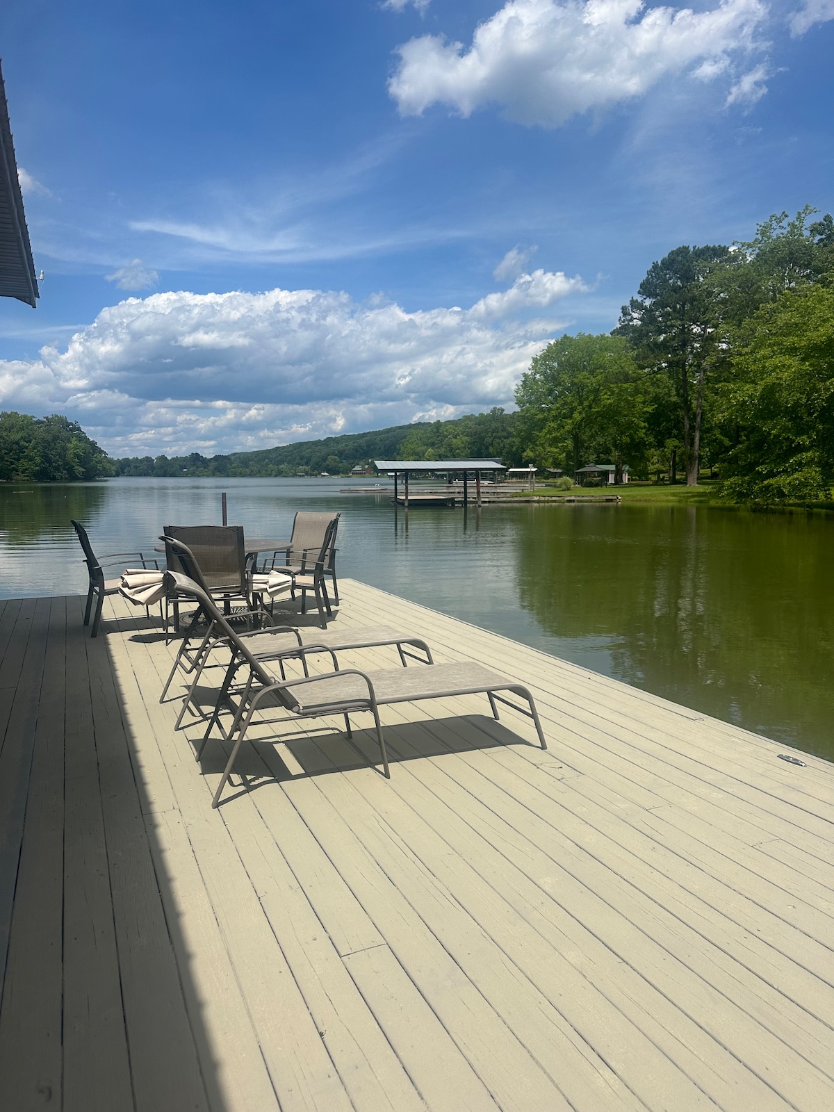 A tranquil dock extends over the calm waters of Watts-Bar Lake, featuring several metal chairs arranged for relaxation. Lush trees line the shore, and the sky is filled with soft clouds, providing a serene backdrop for outdoor enjoyment.