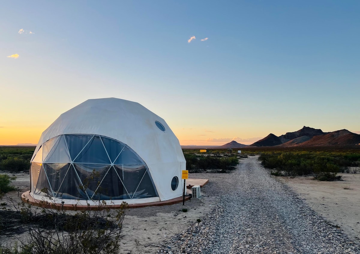 A spacious white dome is presented against a colorful sunset sky, with large triangular windows reflecting the light. A gravel path leads up to the dome, surrounded by desert vegetation, while distant mountain silhouettes create a dramatic backdrop.