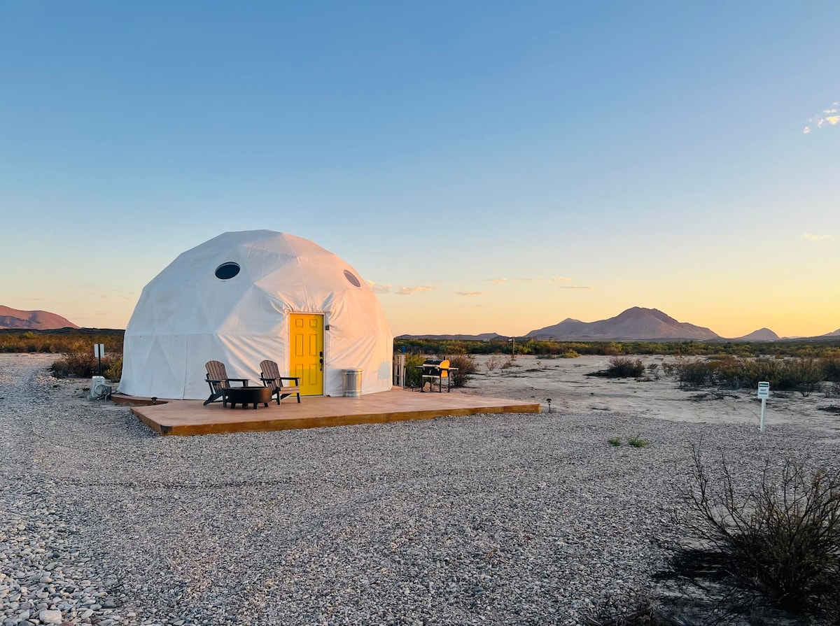 A spacious dome structure is set against a backdrop of desert landscape, featuring a bright yellow door and a private wooden patio. Outdoor seating is provided on the deck, while distant mountains and a colorful sunset create a serene atmosphere.