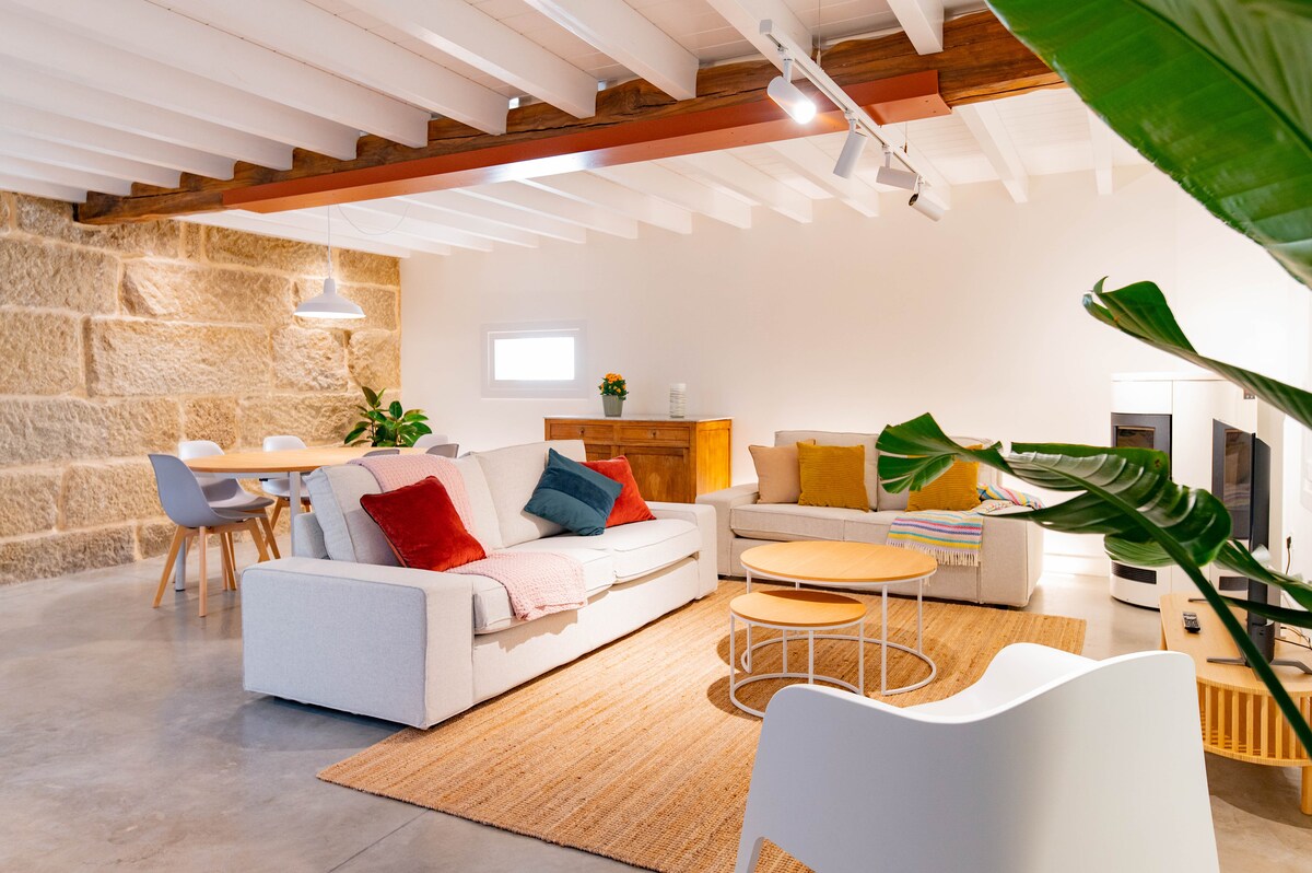 A spacious living area features light-colored sofas with colorful throw pillows, arranged around a circular coffee table. A warm area rug brightens the concrete floor, and a dining table with chairs stands in the background. Natural light streams in from a nearby window.