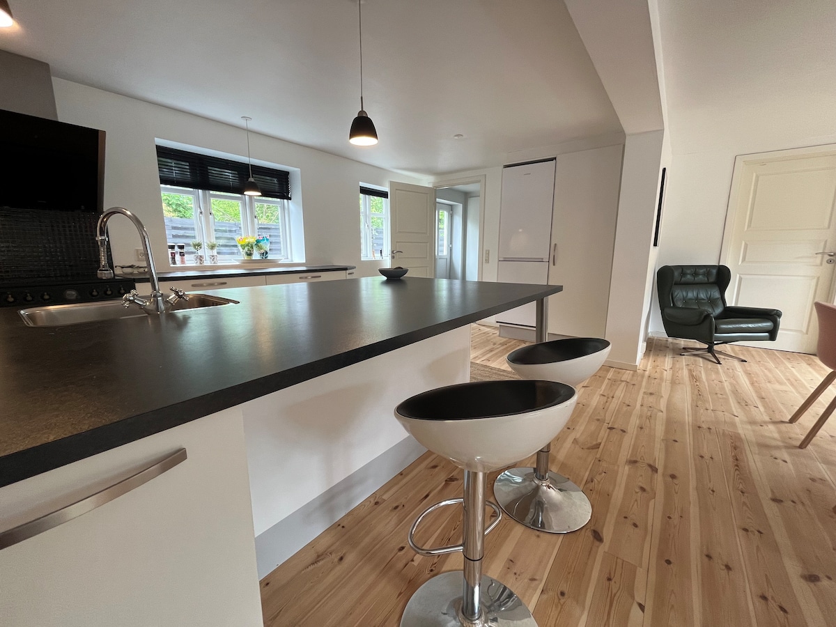 A modern kitchen is shown with stylish bar stools at an island counter. Light wood flooring complements the sleek black countertops, while large windows provide natural light. Minimalist cabinetry and appliances are neatly integrated into the space, enhancing functionality.