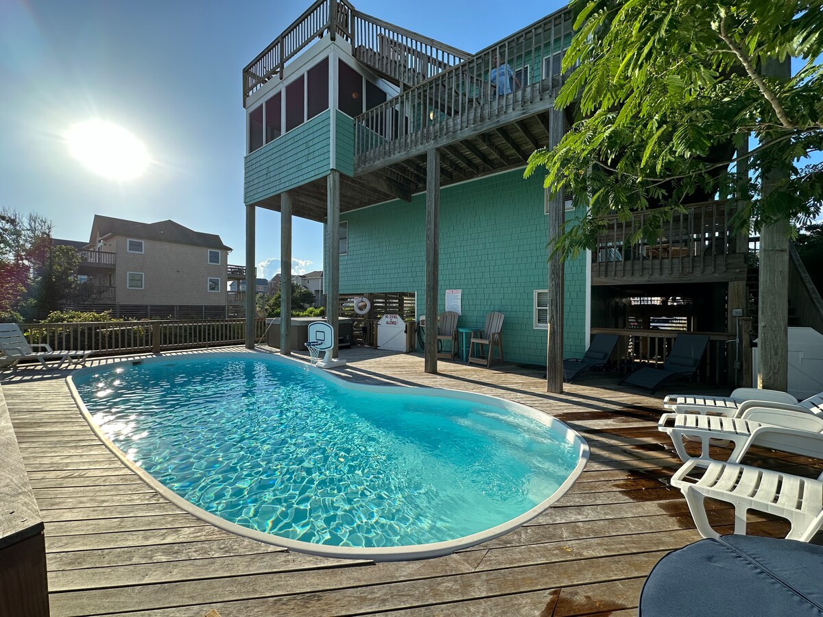 A spacious outdoor area features a large private pool surrounded by wooden decking. Sun loungers are positioned nearby under the shade of trees. The vibrant home stands in the background, showcasing its raised structure and multiple deck levels.