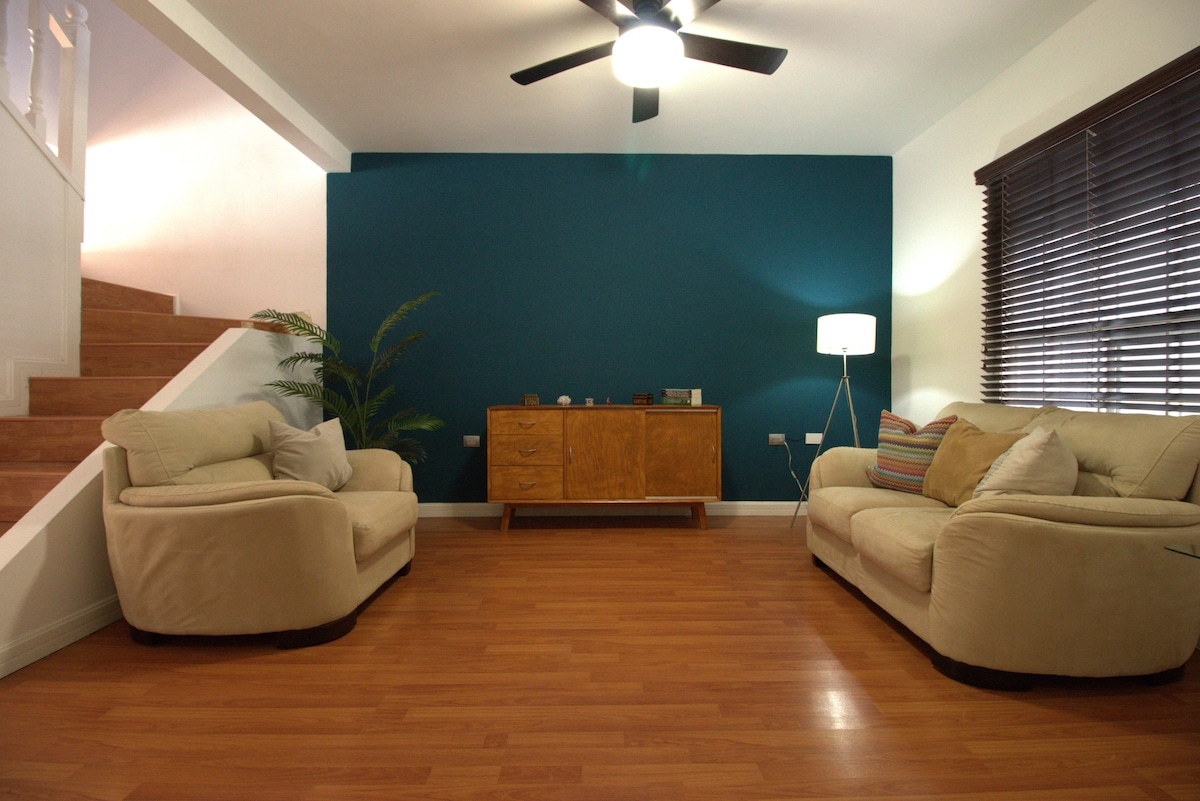 A welcoming living area is adorned with two comfortable sofas facing each other. A wooden sideboard is positioned against a deep teal wall, complemented by a floor lamp. Natural light filters through the dark blinds, and a staircase leads to the upper level.