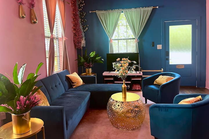غرفة معيشة ساحرة بألوان زرقاء وردية نابضة بالحياة ومقاعد فاخرة ولمسات ذهبية ومساحات خضراء مورقة - مزيج مثالي من الراحة والأناقة لإقامتك.