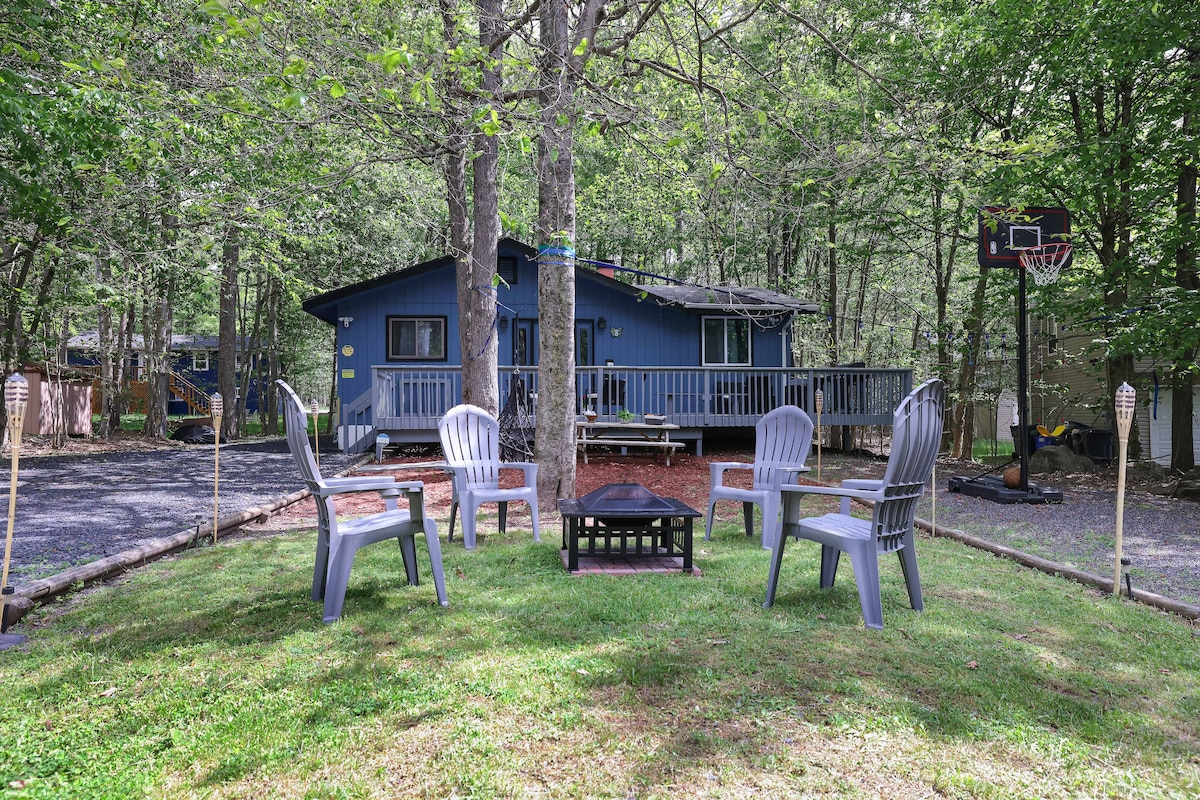 An outdoor space features a grassy area surrounded by trees, with a central fire pit and four chairs arranged around it. The blue house can be seen in the background, accompanied by a basketball hoop and deck area.