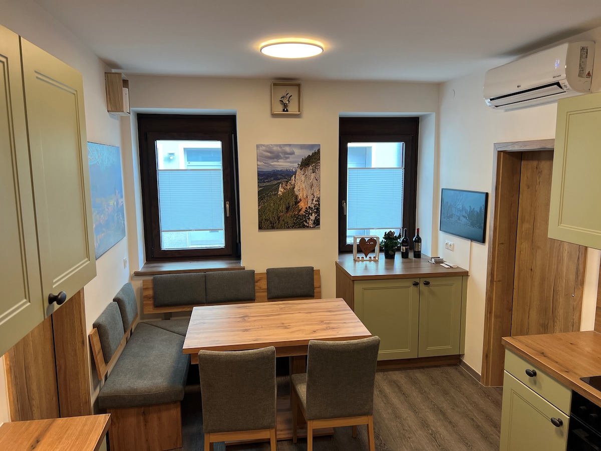 A cozy dining area is presented with a wooden table surrounded by upholstered chairs. Natural light enters through two windows, enhancing the tranquil atmosphere. Wall-mounted artwork and an air conditioning unit add to the space's functionality and comfort.