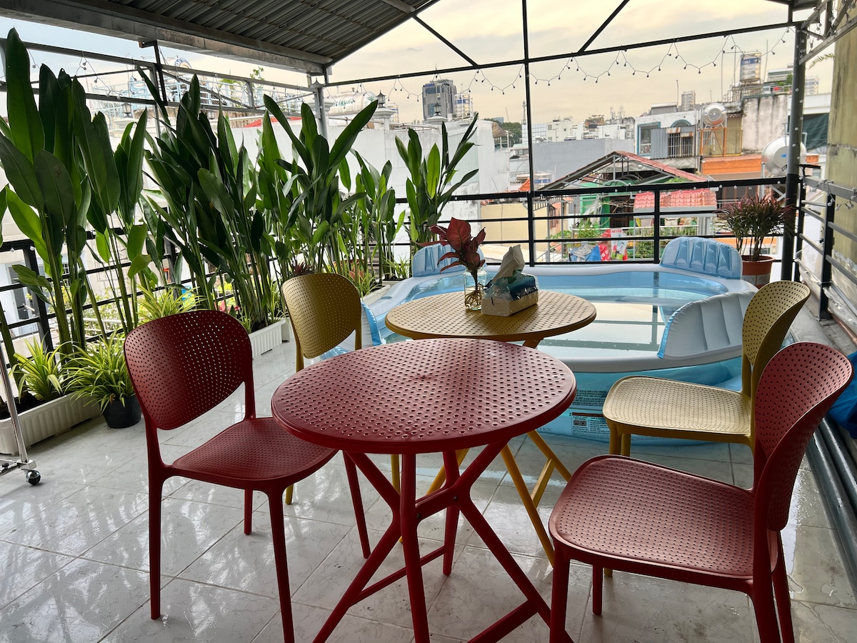 A shared rooftop garden is featured, furnished with a round table and four chairs in red and yellow tones. Potted plants surround the area, providing greenery. A small pool is visible in the background, complemented by city views under a soft sky.