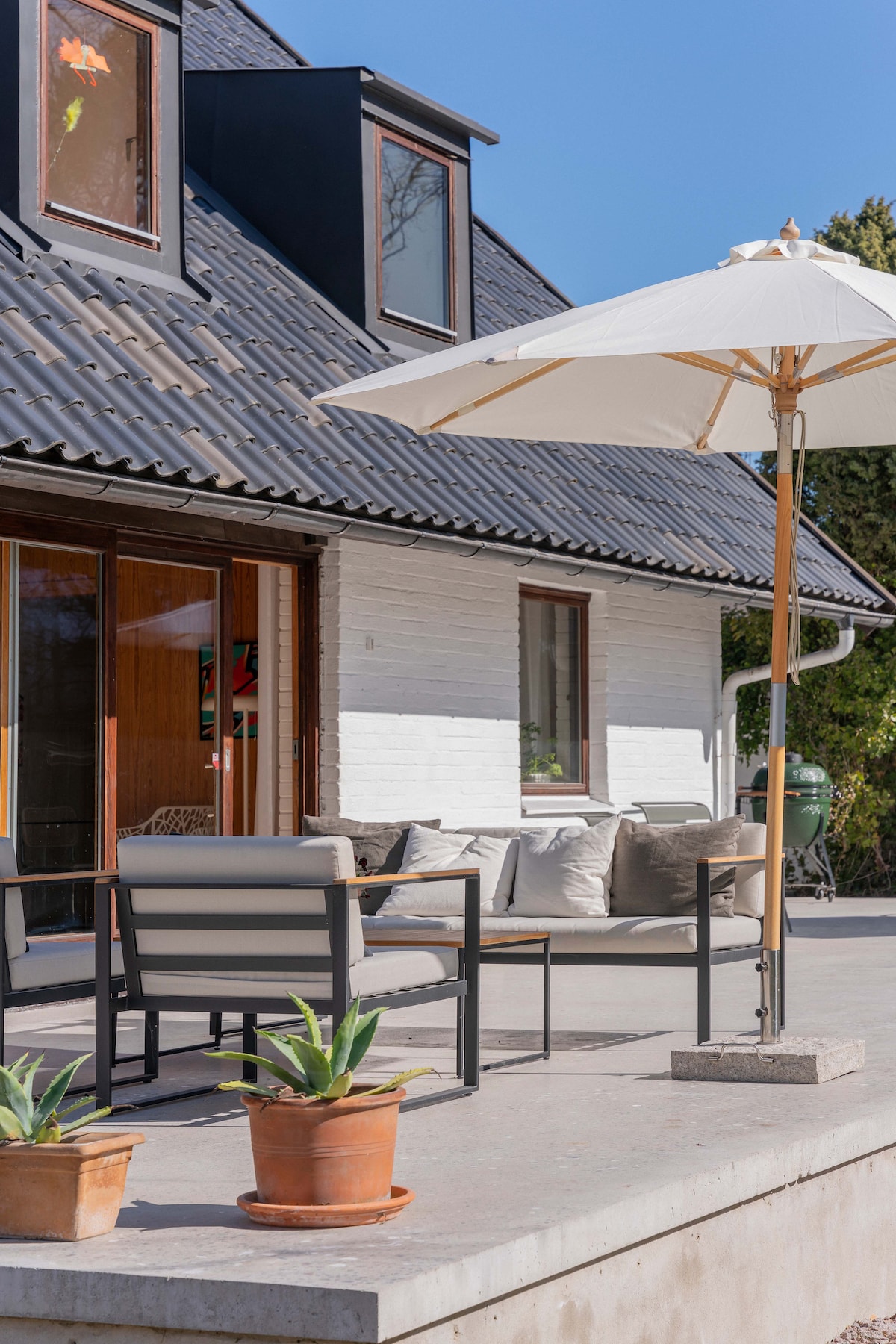 A spacious outdoor area features cozy seating arrangements with cushions, a large white umbrella providing shade, and potted plants. The modern architecture is complemented by large glass doors that open to the inside, enhancing the inviting atmosphere of the home.