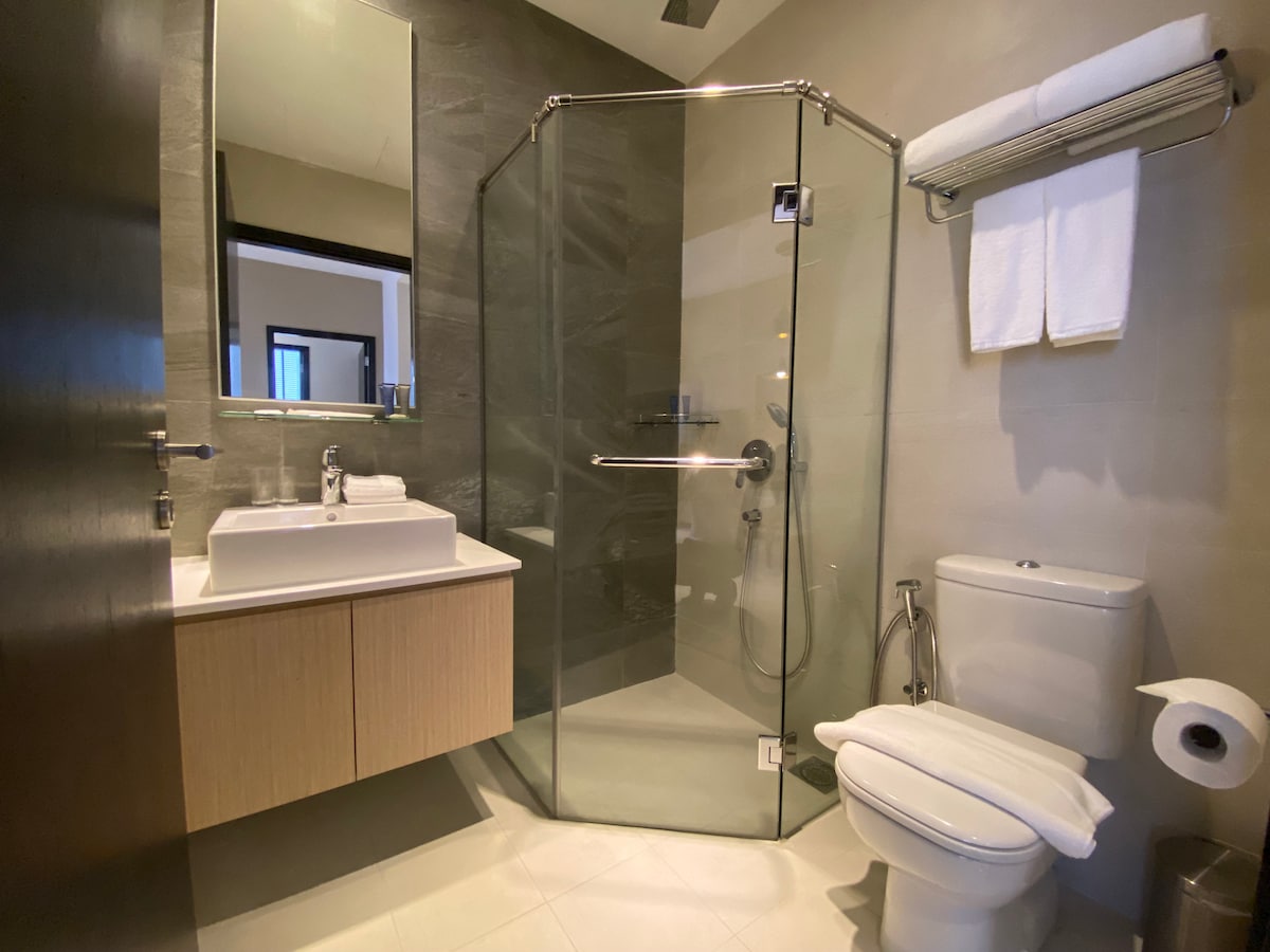 A modern bathroom is showcased, featuring a glass-enclosed shower with a rainfall showerhead. A sleek sink is positioned on a light wood vanity, while white towels are neatly arranged on a wall-mounted bar. A discreet toilet is placed nearby, and minimalistic decor enhances the clean design.