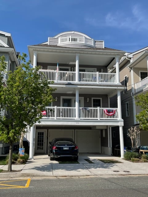 The exterior of a multi-level condominium showcases a classic coastal design with white railings and decorative balconies. A dark-colored vehicle is parked in the garage beneath the building, while colorful bunting decorates the balconies. Lush green landscaping surrounds the entrance.