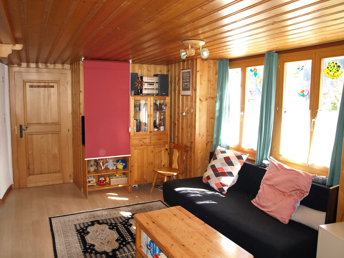 A cozy living area features warm wood paneling and natural light streaming through large windows with light curtains. A black sofa adorned with colorful pillows sits adjacent to a wooden coffee table. A bookshelf displays various items, contributing to the inviting atmosphere.