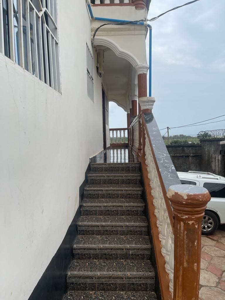 An exterior view highlights a staircase leading to an upper level. The stairs feature a textured surface, and the railing is crafted from polished wood, providing a contrast against the white walls of the building. A cloudy sky is visible in the background.
