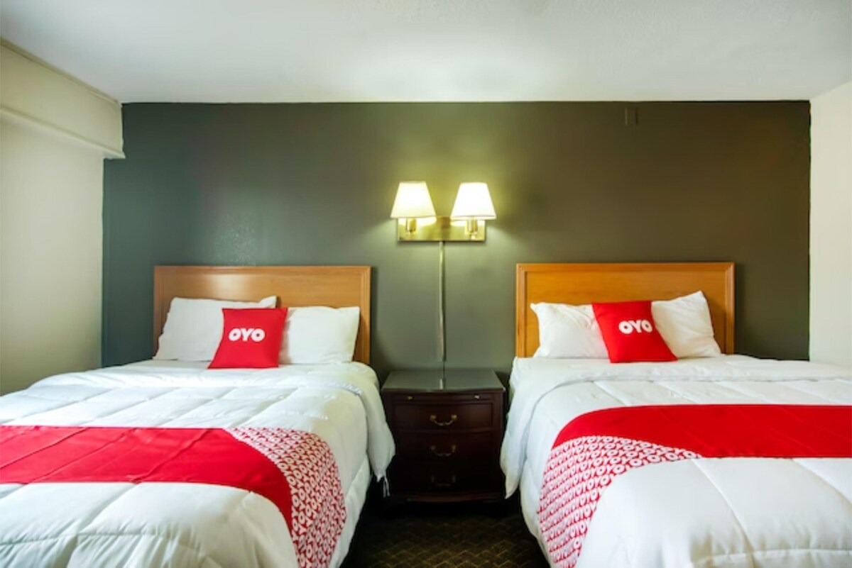 A well-lit sleeping area features two spacious beds with white bedding and vibrant red accents. Nightstands with lamps are placed between the beds. The wall behind is painted a dark color, providing a modern contrast to the light linens.