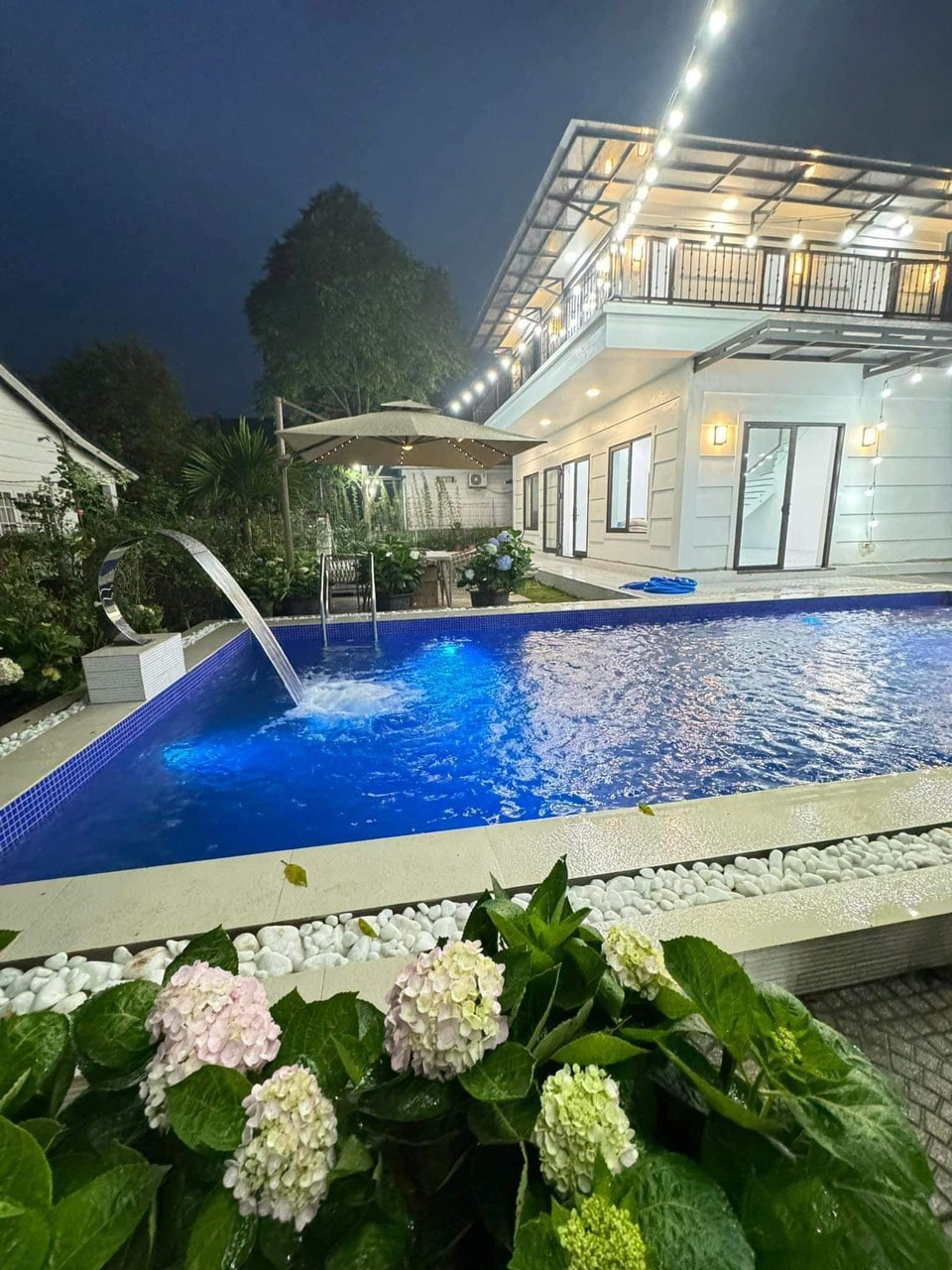 A tranquil pool area features a shimmering blue pool with a sleek water feature. Surrounding greenery and blooming flowers enhance the setting, while a spacious building with glass doors and outdoor seating is visible in the background, softly illuminated by warm lights.