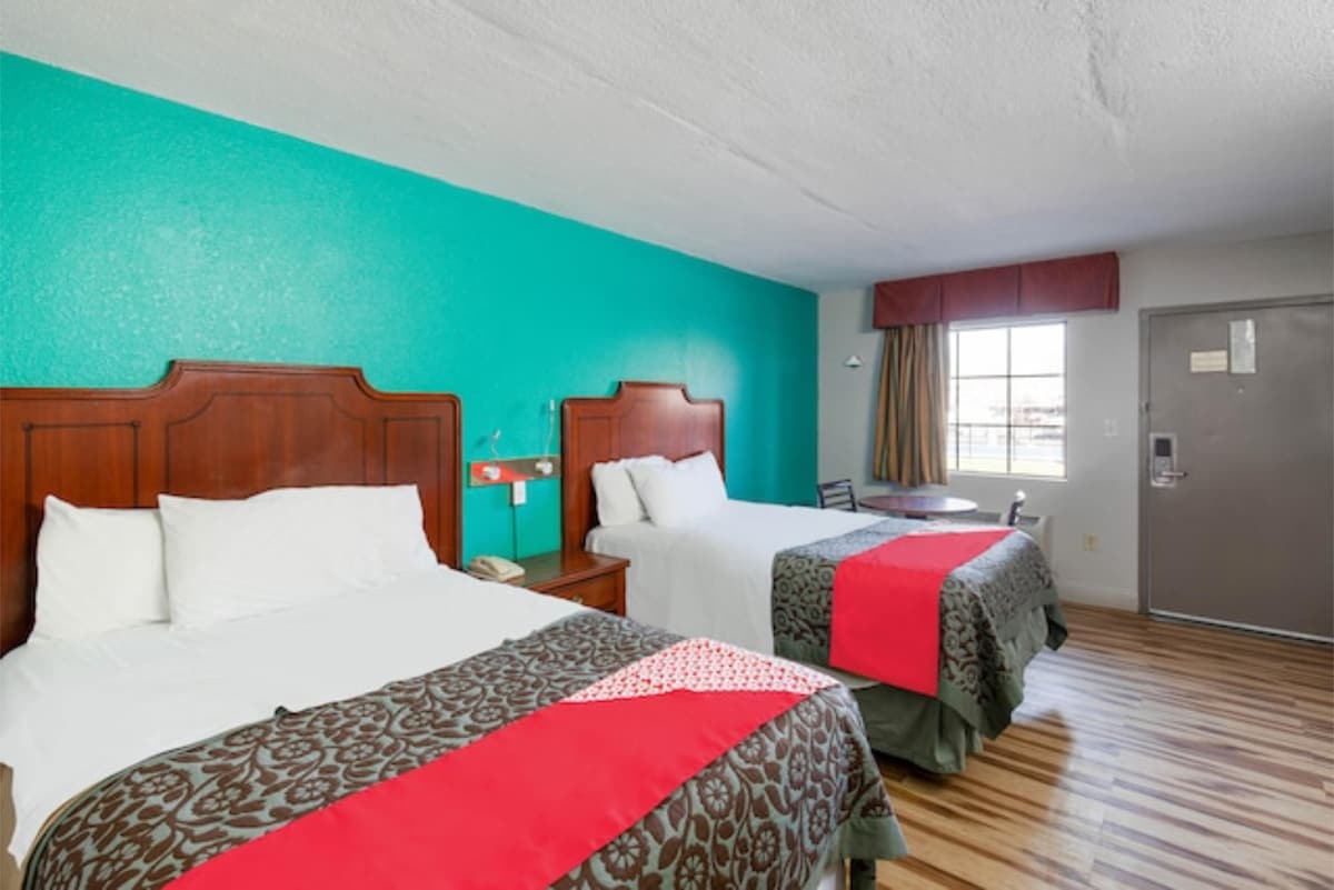 A bright room features two spacious beds with decorative blankets, complemented by a vibrant teal accent wall. Windows allow natural light to fill the space, and a small table with chairs is positioned beside the window, offering a cozy area for guests.