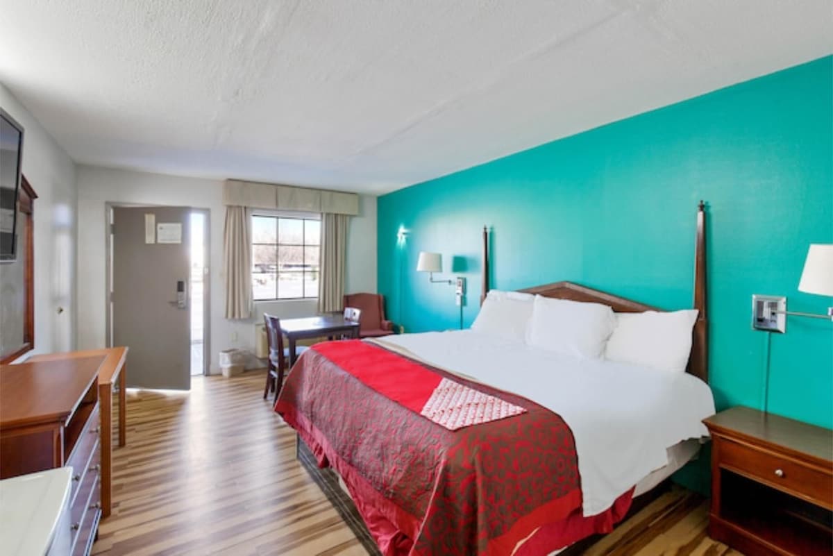 A modern guest room features a spacious bed with crisp white linens, accented by a red blanket. A desk and chair are positioned near a window, which is dressed with light-colored curtains. The walls are painted a vibrant teal, enhancing the contemporary design.
