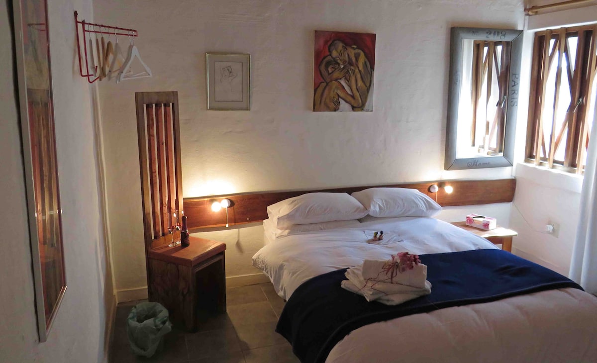 A comfortable sleeping area features a spacious bed with white linens and a dark blanket. Wooden accents are present in the headboard and furniture, while framed artwork adorns the walls. Natural light enters through the windows, partially covered by wooden slats.