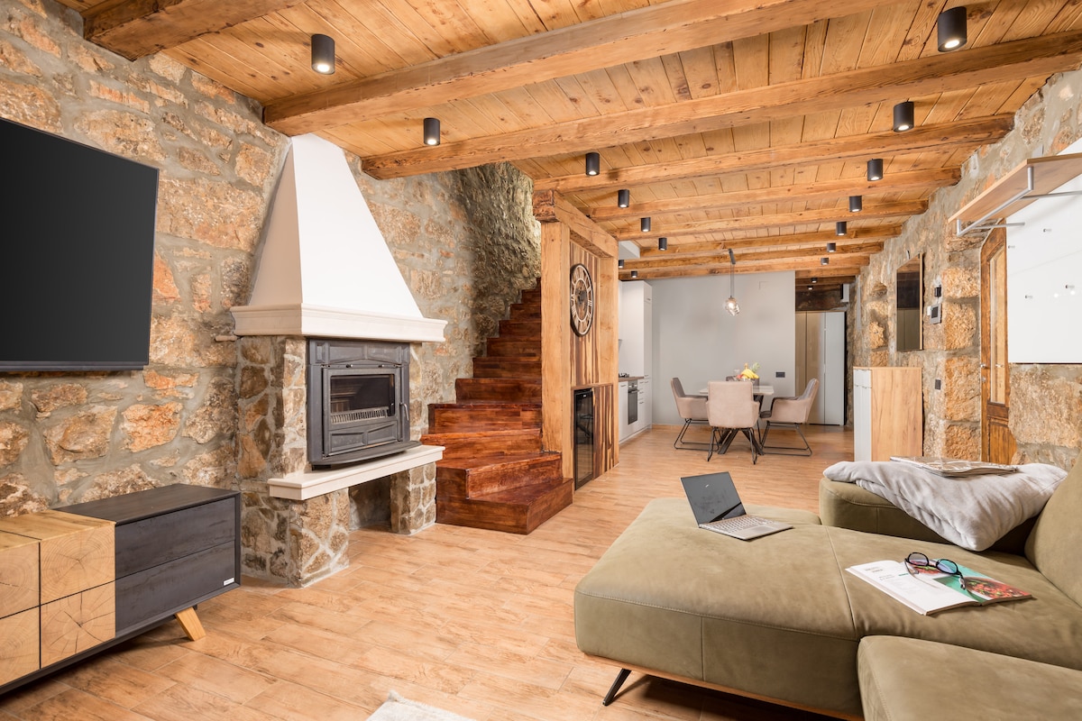 The spacious living area features a stone fireplace and wooden ceiling beams. A corner sofa is complemented by a coffee table with a magazine. An open staircase leads to the upper level, while a dining area is visible in the background with a modern light fixture above.