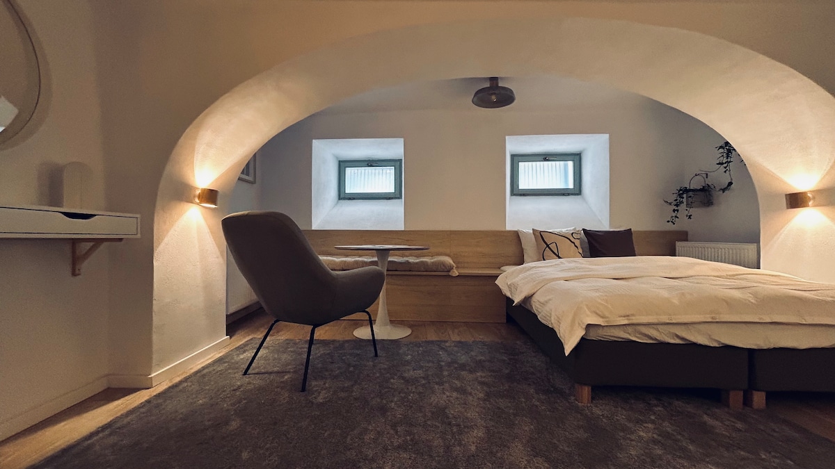 A spacious sleeping area is defined by an arched ceiling, featuring subtle lighting fixtures. A comfortable bed with soft linens is placed against a wooden wall, complemented by a small table and chair nearby. Two windows allow natural light to brighten the space.