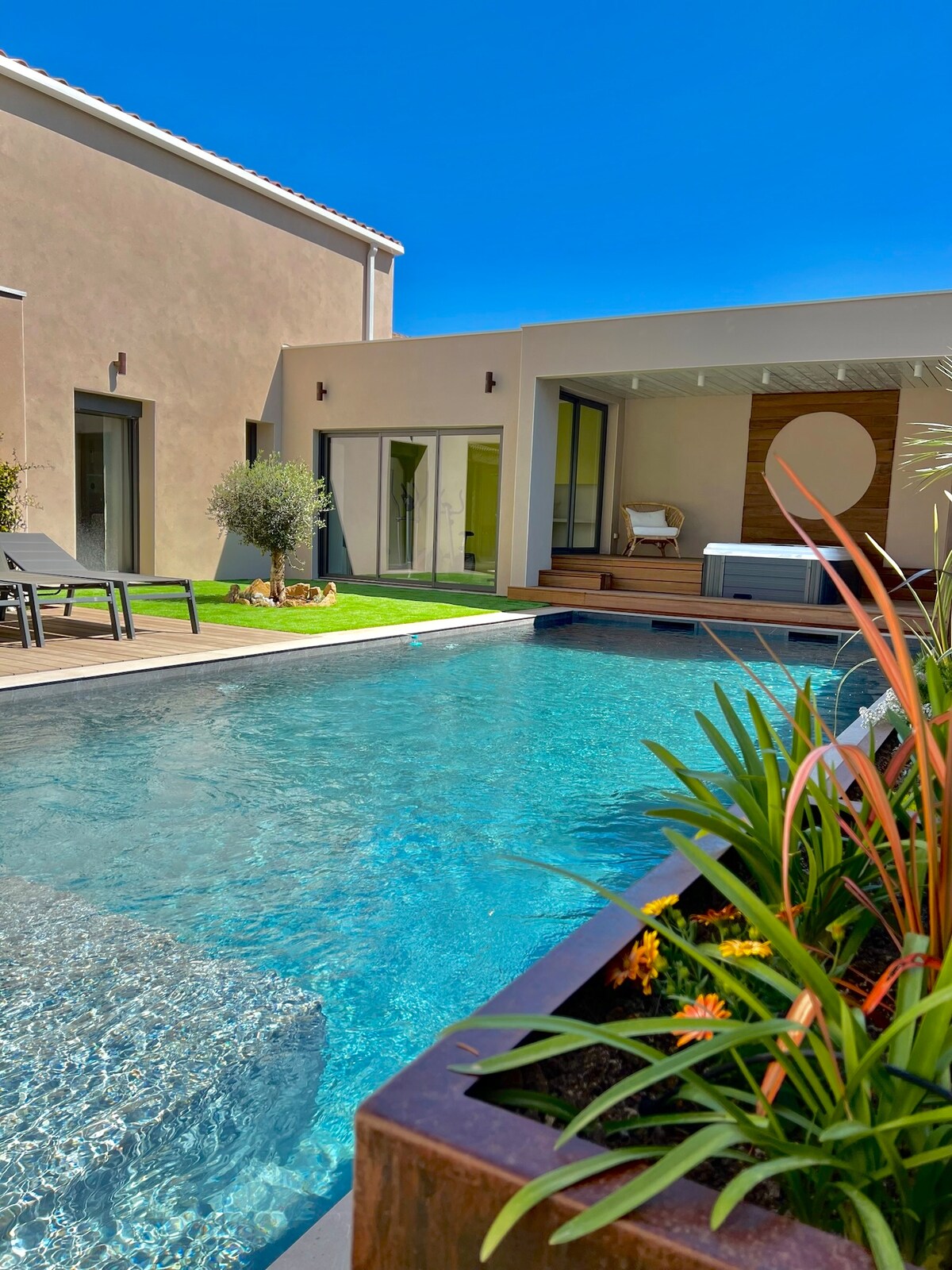 A serene outdoor space features a clear blue pool with a gentle ripple. Modern loungers are positioned on the wooden deck, surrounded by lush greenery. An outdoor spa is partially visible under a covered area, contributing to a relaxing environment.