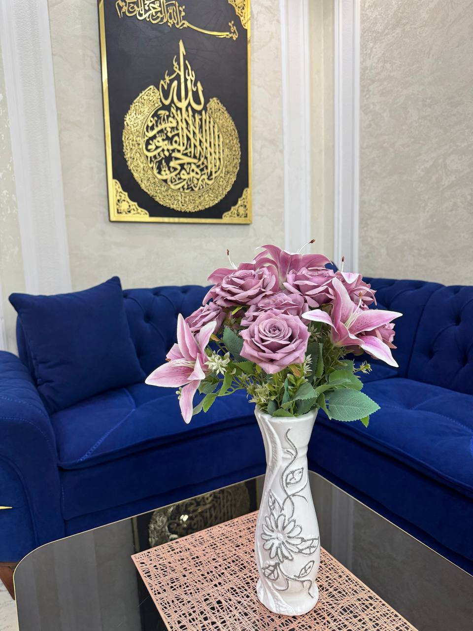 A white vase with floral designs holds a bouquet of pink roses and lilies. Behind it, a deep blue sofa complements a textured wall. A decorative piece featuring intricate calligraphy hangs above, enhancing the room's elegant ambiance.