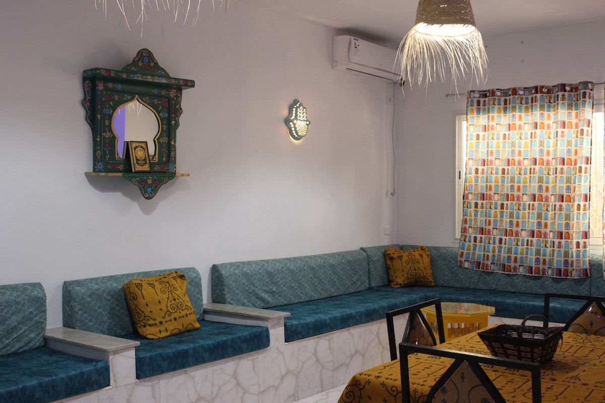 A spacious seating area is designed with built-in benches covered in teal upholstery. Brightly patterned curtains frame the window, allowing natural light to brighten the room. A decorative wall shelf and soft ambient lighting enhance the cozy atmosphere.