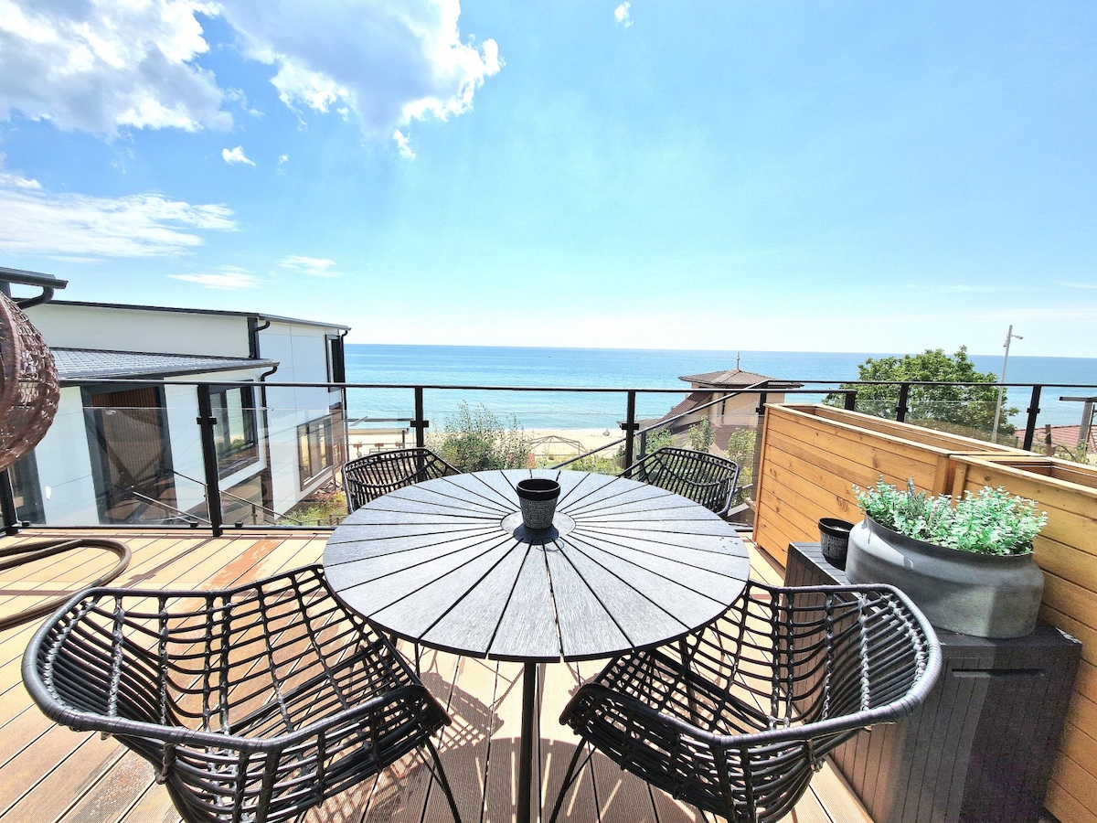 A spacious outdoor terrace is featured, offering a round table surrounded by four black woven chairs. The view includes the calming sea and beach, with clear blue skies and gentle clouds visible in the background.