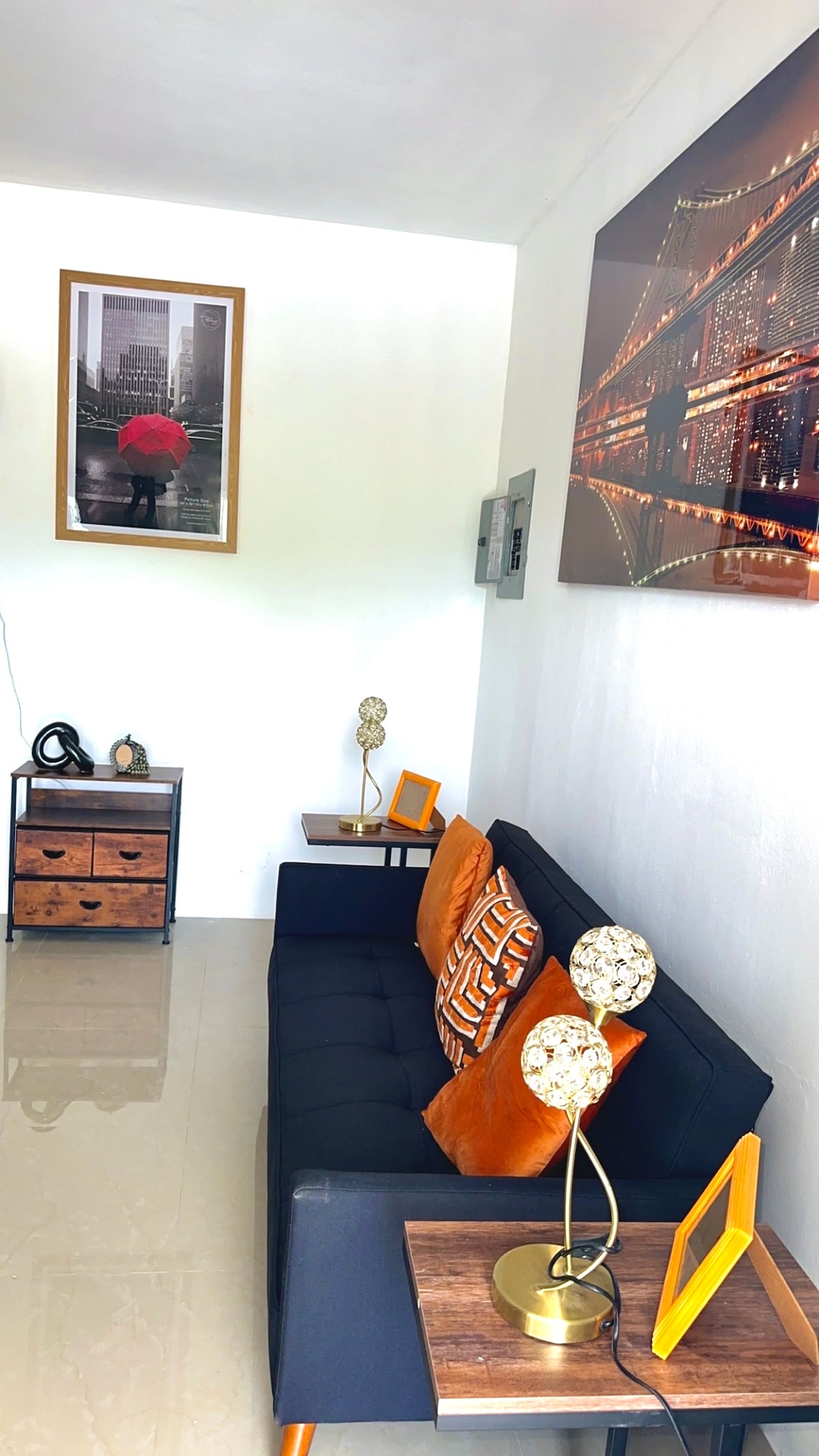 A well-lit sitting area features a stylish black sofa adorned with patterned cushions. Decorative lamps with geometric designs are placed on wooden side tables. A framed photograph of a city scene hangs on the wall, complementing the modern décor and polished tiles.