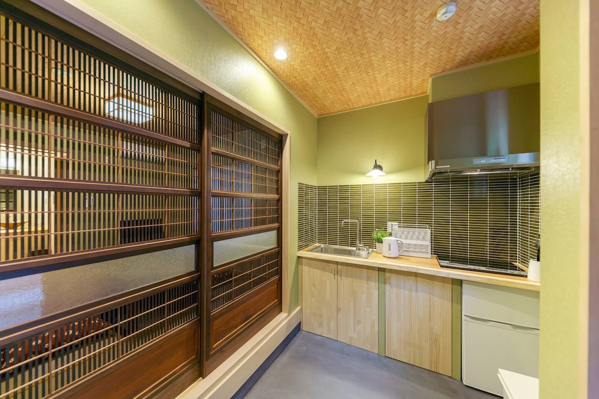 The kitchen features natural wood cabinetry and a counter with a sink and stovetop. Sliding glass doors provide access to storage, adding to the spacious feel. A modern refrigerator stands to the right, with soft lighting enhancing the area’s tranquility.