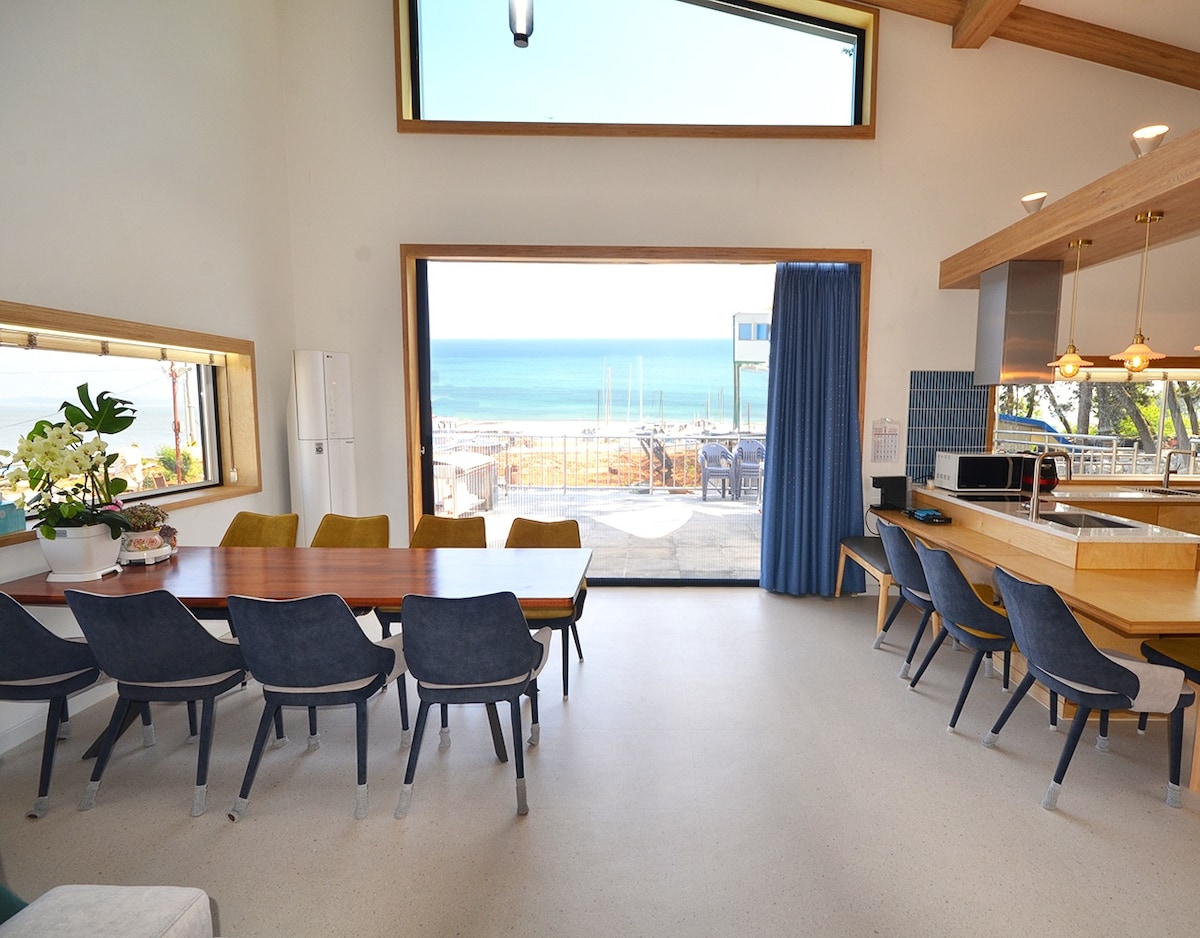 A spacious dining area features a long wooden table surrounded by navy blue chairs. Large windows offer expansive ocean views, with natural light illuminating the space. A modern kitchen area can be seen to the right, enhancing the open concept layout.