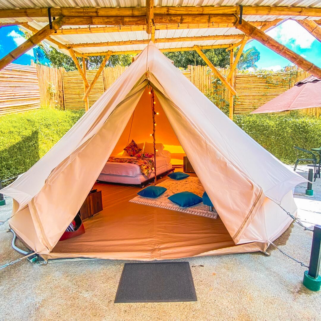 A spacious yurt tent is showcased under a wooden pergola. Inside, two beds can be seen along with decorative pillows placed on the floor. Natural light illuminates the interior, creating a warm ambiance. The outdoor area features surrounding greenery and shaded seating.