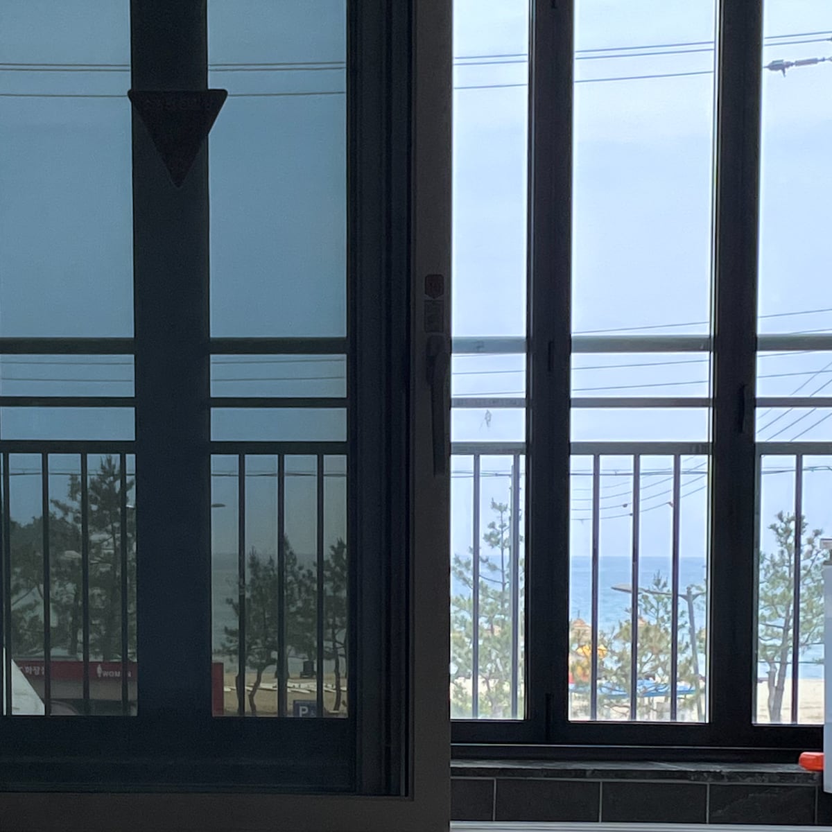 Large glass doors allow natural light to fill the space, revealing a view of the serene outdoor area and the ocean beyond. The balcony is framed by a sleek railing, offering an unobstructed sight of the distant waves and beach trees.