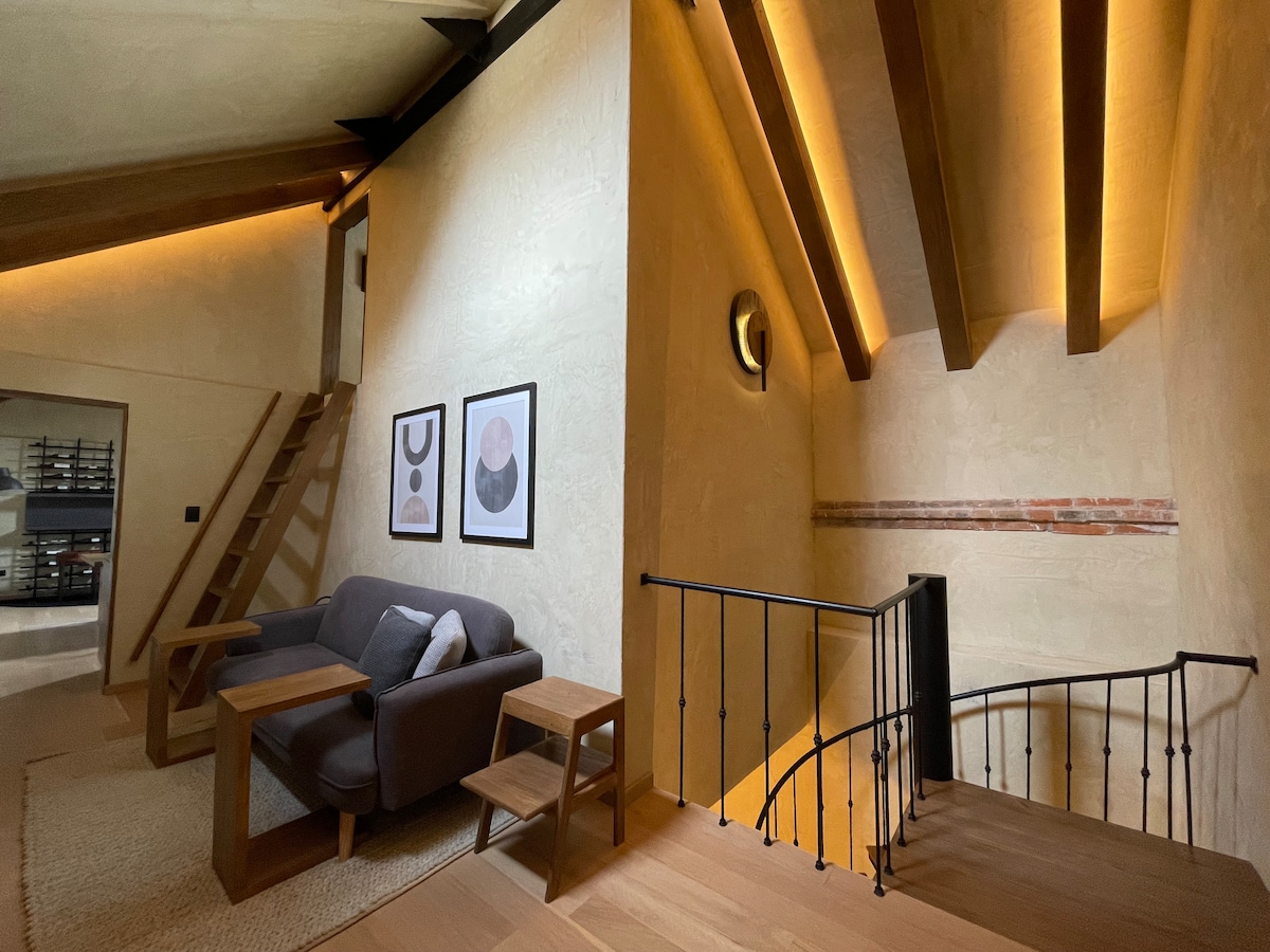 A well-lit corner of the interior features a modern seating arrangement with a dark couch and wooden side tables. A staircase leads to an upper level, and soft lighting highlights the textured walls, complemented by framed artwork displaying abstract designs.