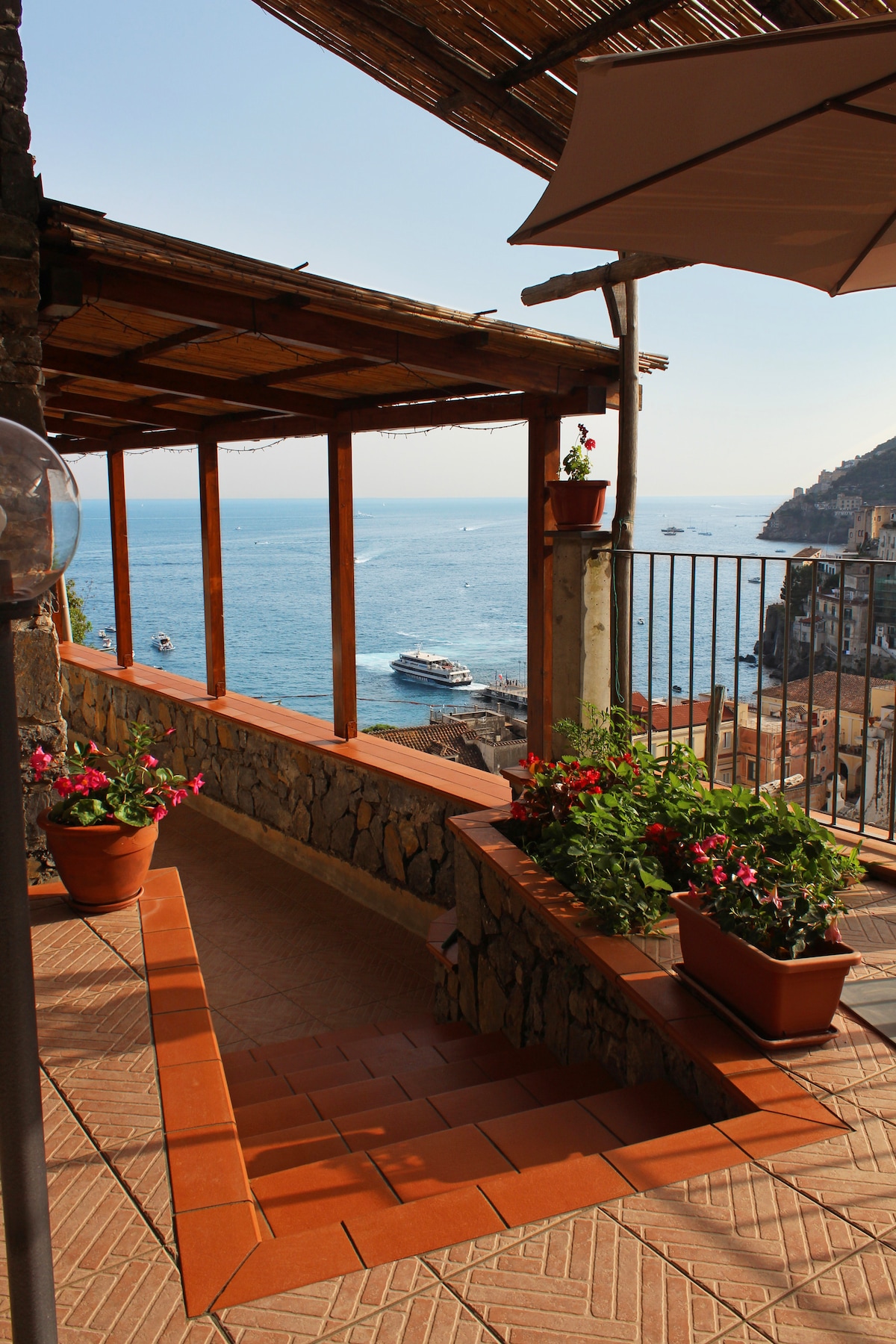 An outdoor terrace is featured, framed by wooden beams and shaded by a fabric awning. Potted flowers in vibrant colors enhance the scene, while a view of the sparkling sea and distant boats creates a serene atmosphere. Stone walls border the pathway leading to the terrace.