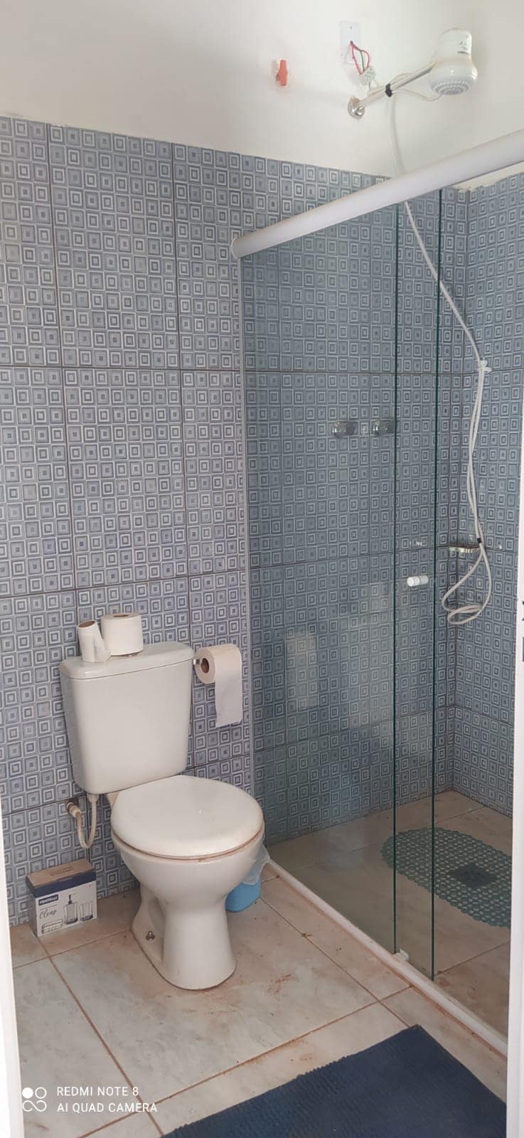 A well-appointed bathroom features a glass shower enclosure alongside a white toilet. The walls are adorned with a patterned blue tile, which enhances the clean appearance. A small mat is placed on the floor for comfort, and essential toiletries are neatly arranged.