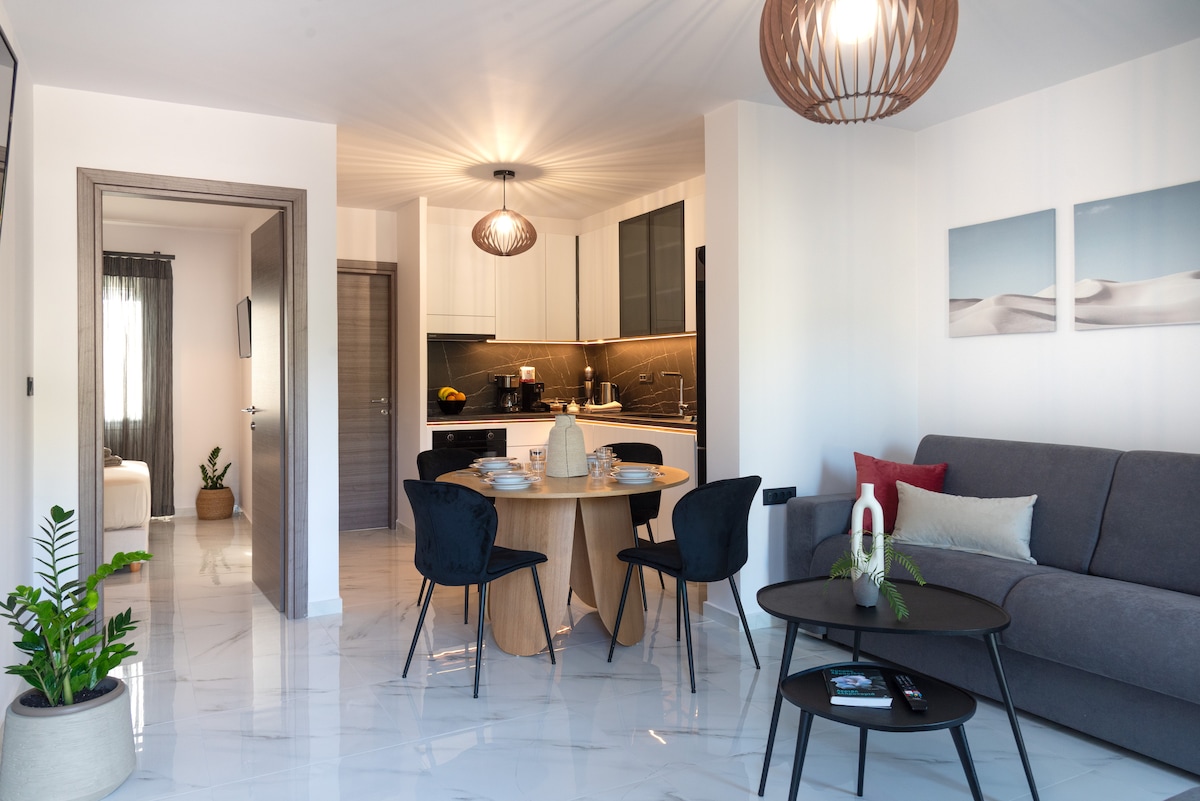 An inviting living space is presented, featuring a round dining table set for four, surrounded by black chairs. A gray sofa offers comfortable seating, with a low coffee table nearby. The open kitchen area showcases modern cabinetry and dining essentials, with soft lighting overhead enhancing the ambiance.